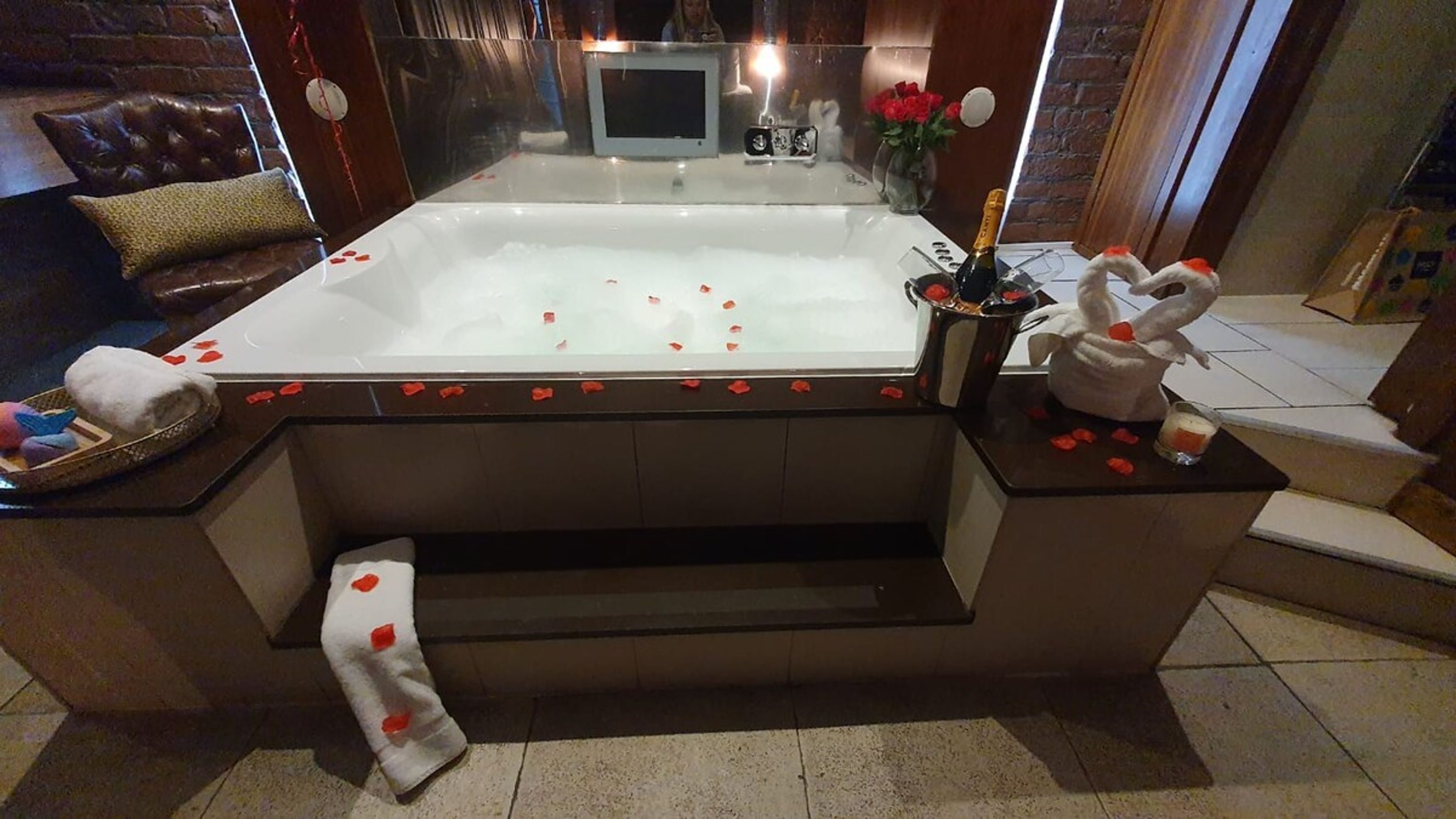 Un grand bain à remous rectangulaire, dont la surface est parsemée de pétales de rose, entouré d’un banc de couleur sombre et sur lequel est drapée une serviette.