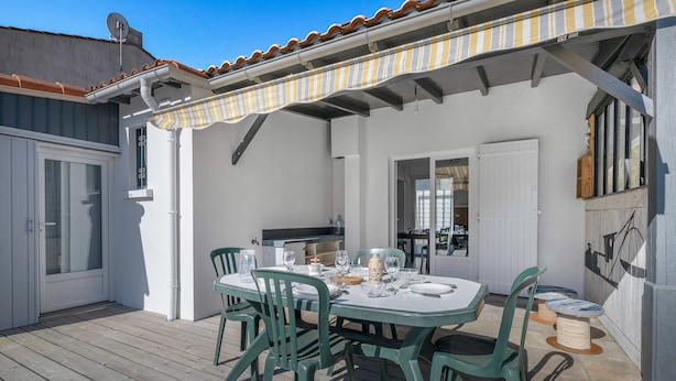 A covered outdoor dining area with a table set for four, surrounded by green chairs, and a kitchen visible through an open door. - 300m from the sea - garden and small pool (Pornic)