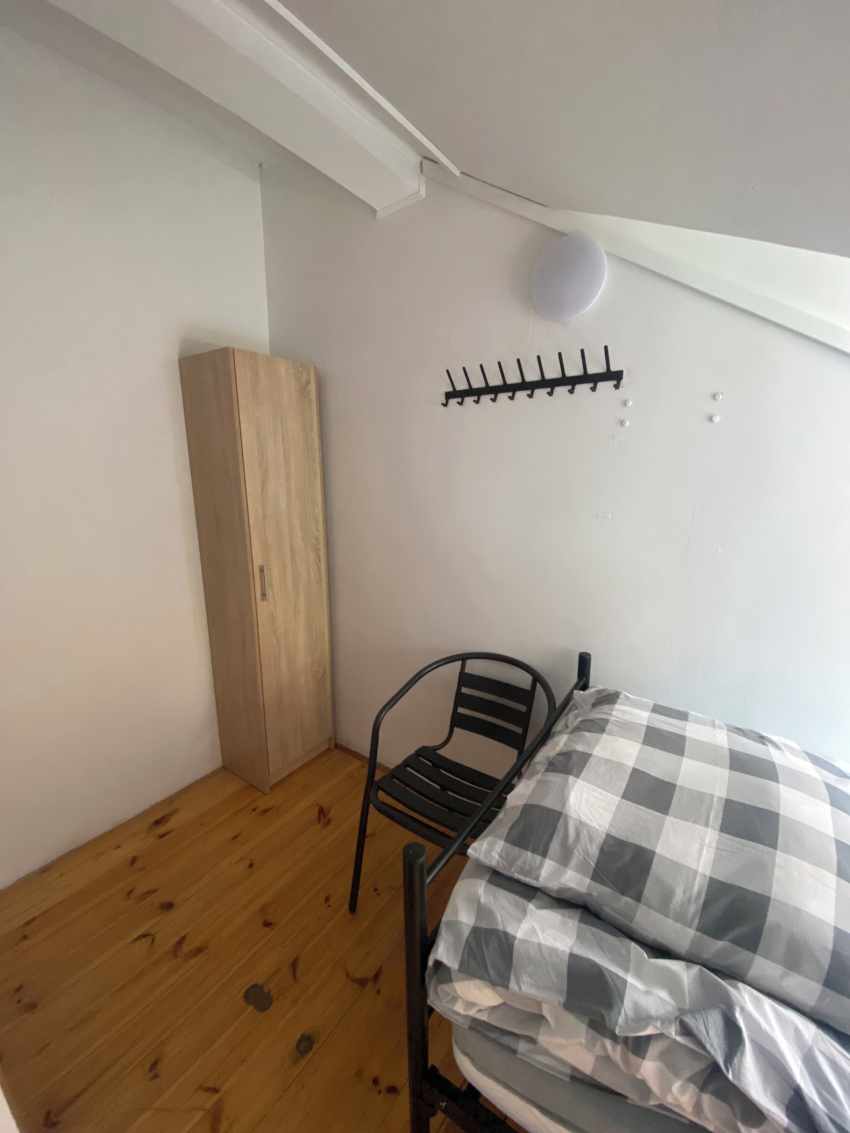 Une petite pièce, peu meublée, avec une armoire en bois, une chaise en métal et un lit recouvert d’une couette à carreaux.
