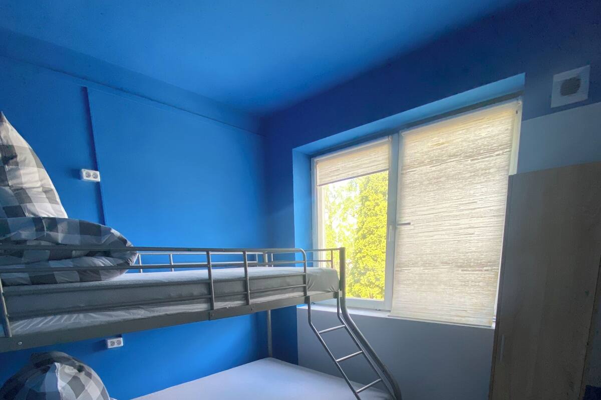 A dormitory room with two bunk beds, a window with blinds, a chair, and a ceiling light.