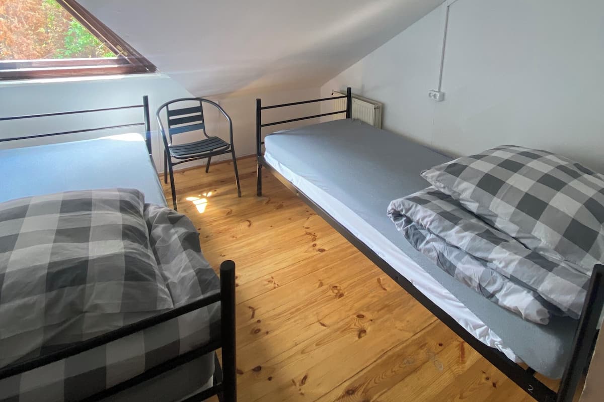 A small, single-room with a sloped ceiling, two metal bunk beds, a wooden floor, a chair, and a window providing natural light.