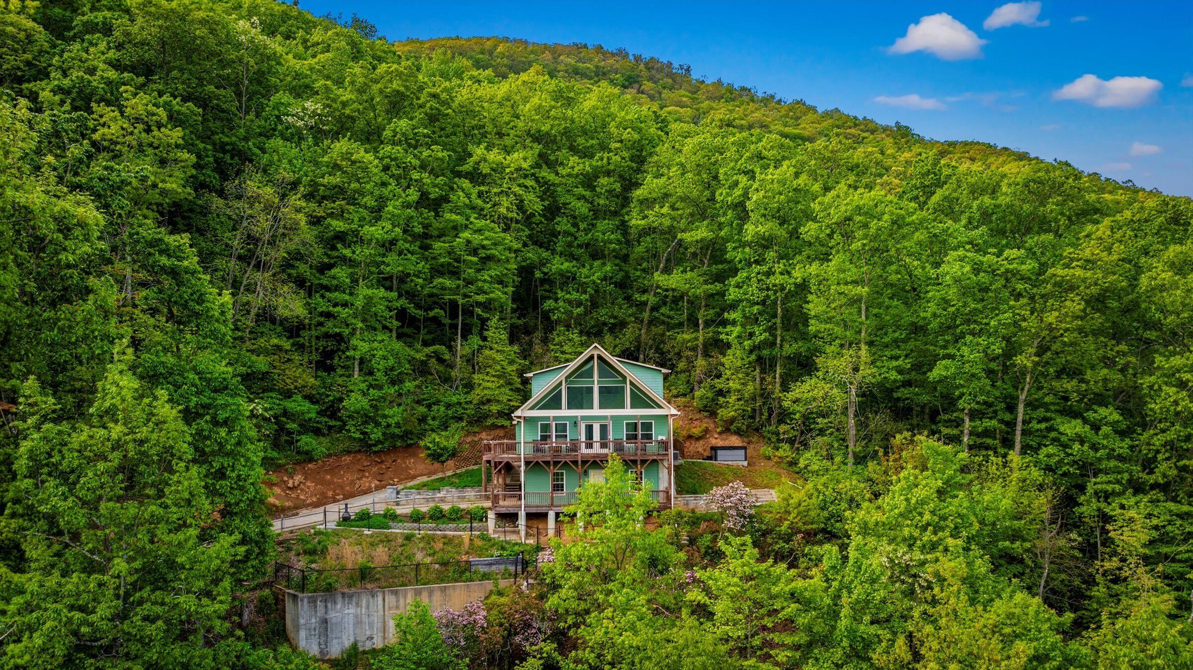 A large, two-story house with a prominent front porch and a wrap-around deck, surrounded by dense forest.