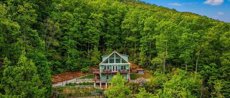 A large, two-story house with a prominent front porch and a wrap-around deck, surrounded by dense forest.