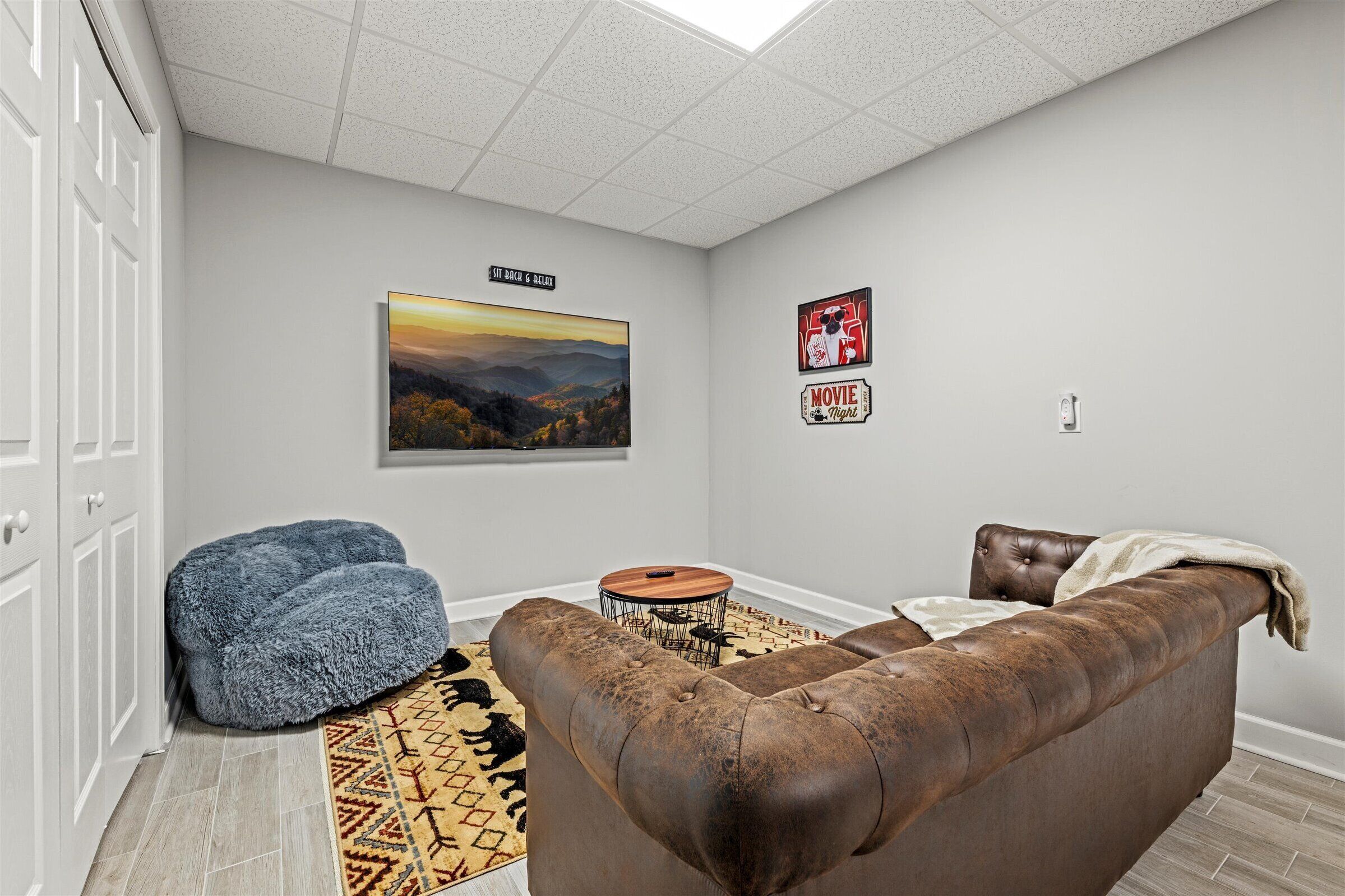 A living room with a brown leather sofa, a blue pouf, a round coffee table, a patterned rug, and a framed landscape painting.