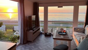 Ein Zimmer mit großem Fenster mit Blick auf einen Strand bei Sonnenuntergang, einem Fernseher, einer Couch und einem kleinen Tisch mit Getränken.