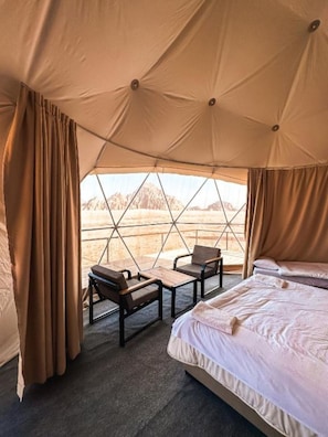 A tented room with two beds, a table, and chairs, with a view of mountains through the window. - Moonlight Camp (Wadi Rum)
