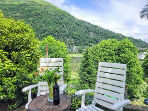 A table with a bottle and flowers, two white chairs, and a view of a lush green valley with a mountain in the background. - Nice apartment for 3 people with WIFI, TV, balcony, pets allowed and panoramic view (Losone)