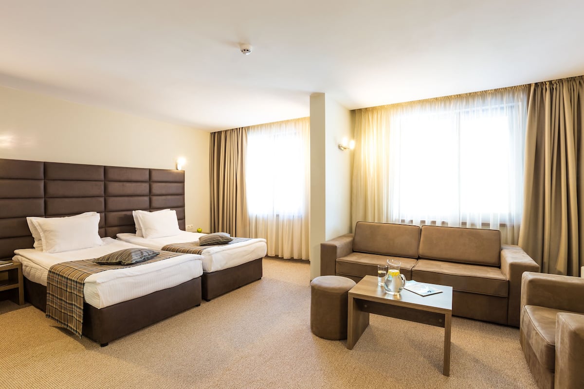 A hotel room with two beds, a sofa, a coffee table, and large windows with curtains.