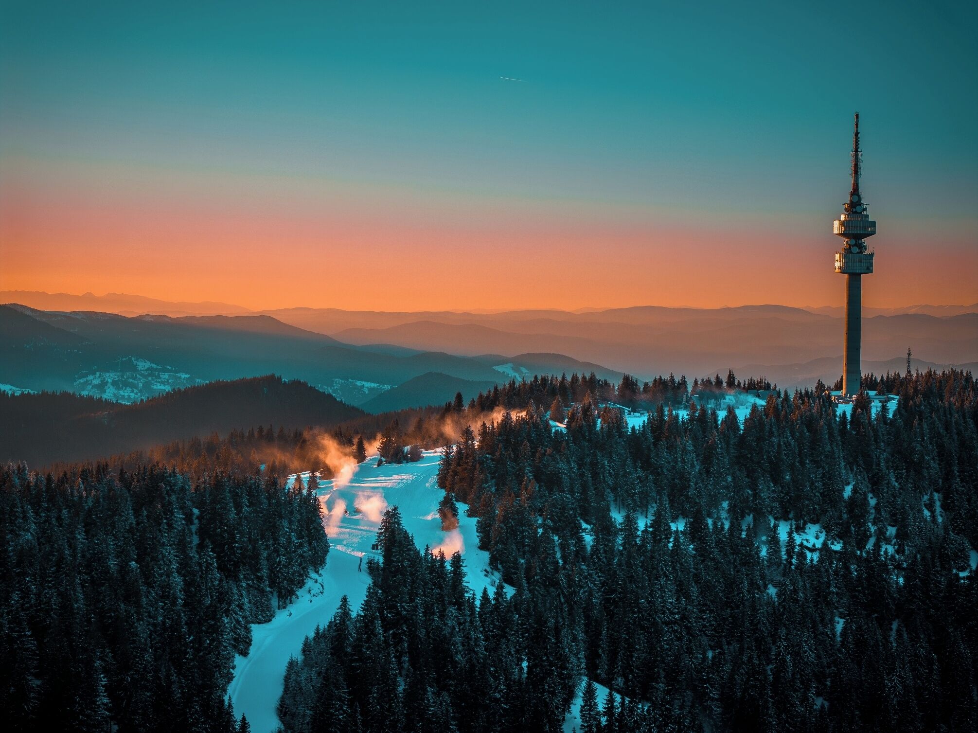 Ein weiter Blick von erhöhter Position auf ein Gebirge mit einem markanten Turm, umgeben von dichten Wäldern und schneebedeckter Landschaft.