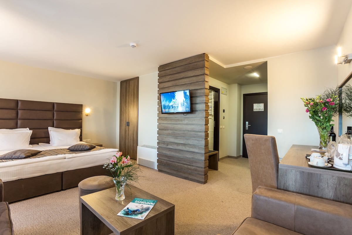 A modern hotel room with a large bed, a flat-screen TV mounted on a wooden panel, a coffee table with a magazine, and a dining area with a vase of flowers.