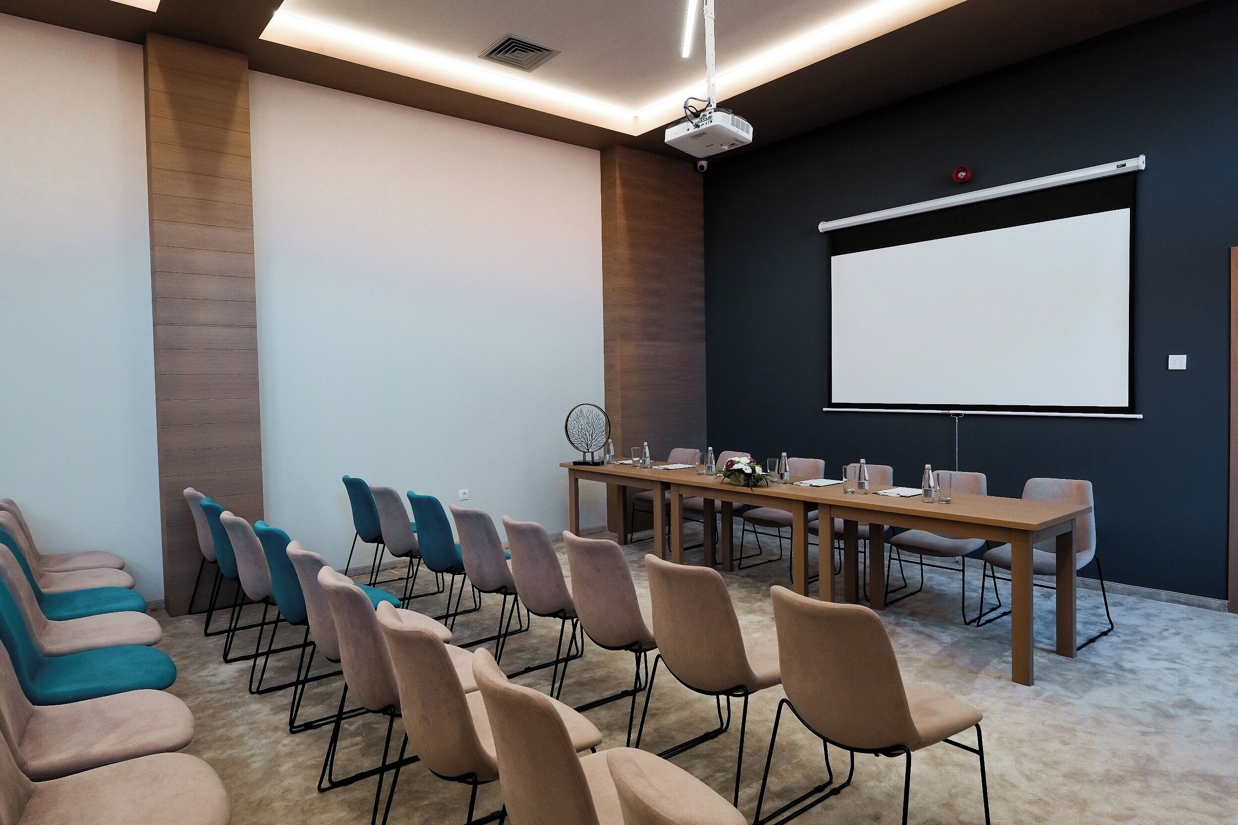 Un salón de conferencias con una larga mesa, sillas, una pantalla de proyector y una pizarra.