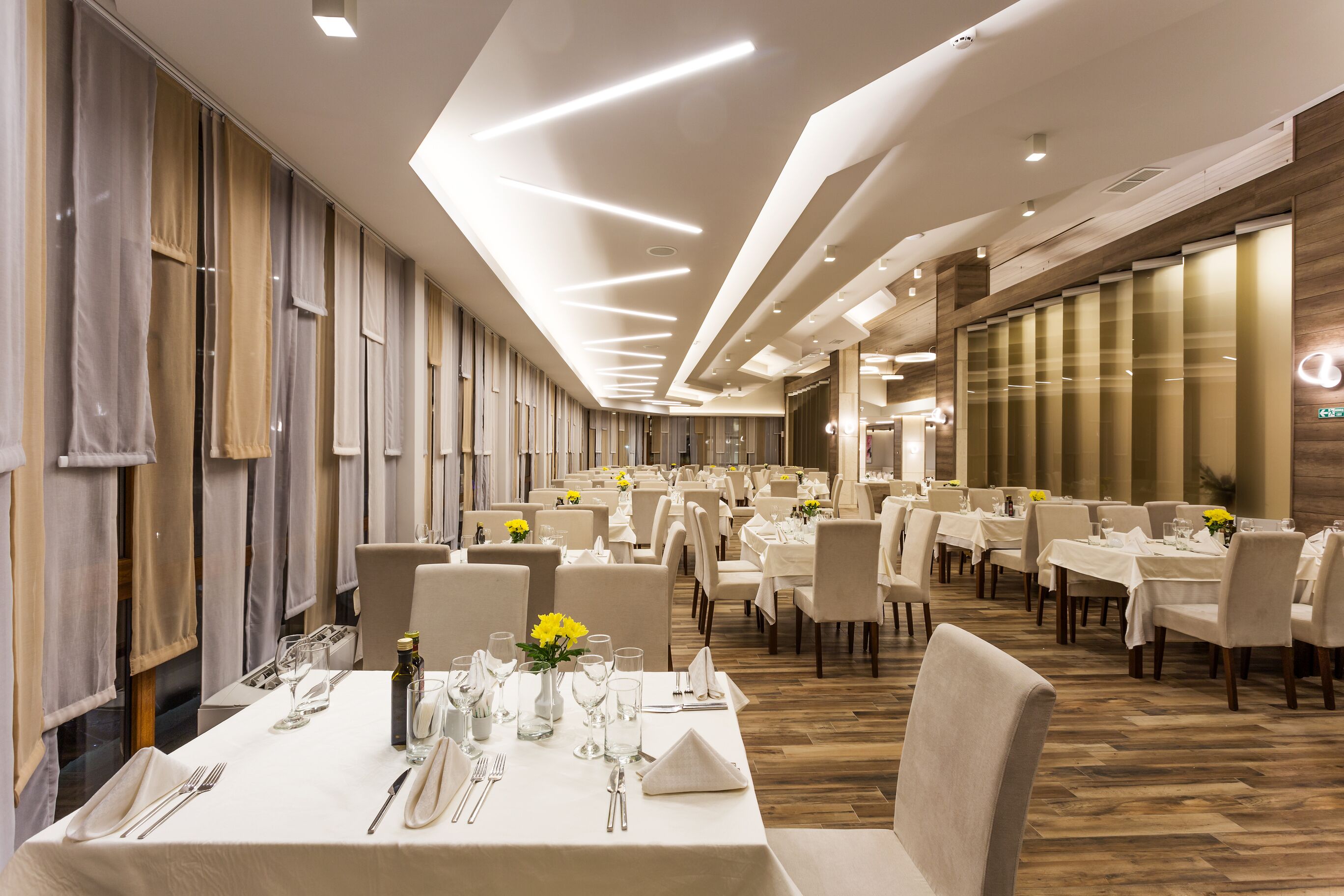 A spacious restaurant with multiple tables set for dining, featuring a modern ceiling design and wooden paneling on the walls.