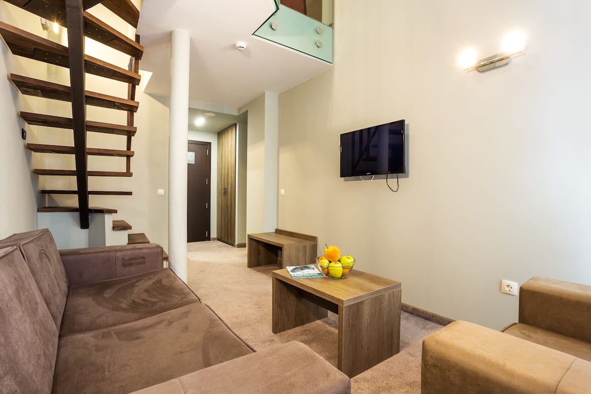 A modern living room with a brown sofa, a wooden coffee table with fruit, and a TV mounted on the wall.