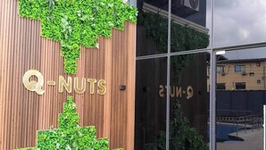 Entrada de un edificio con pared de madera y follaje verde, donde se muestra el nombre "Q-NUTS" de forma destacada.