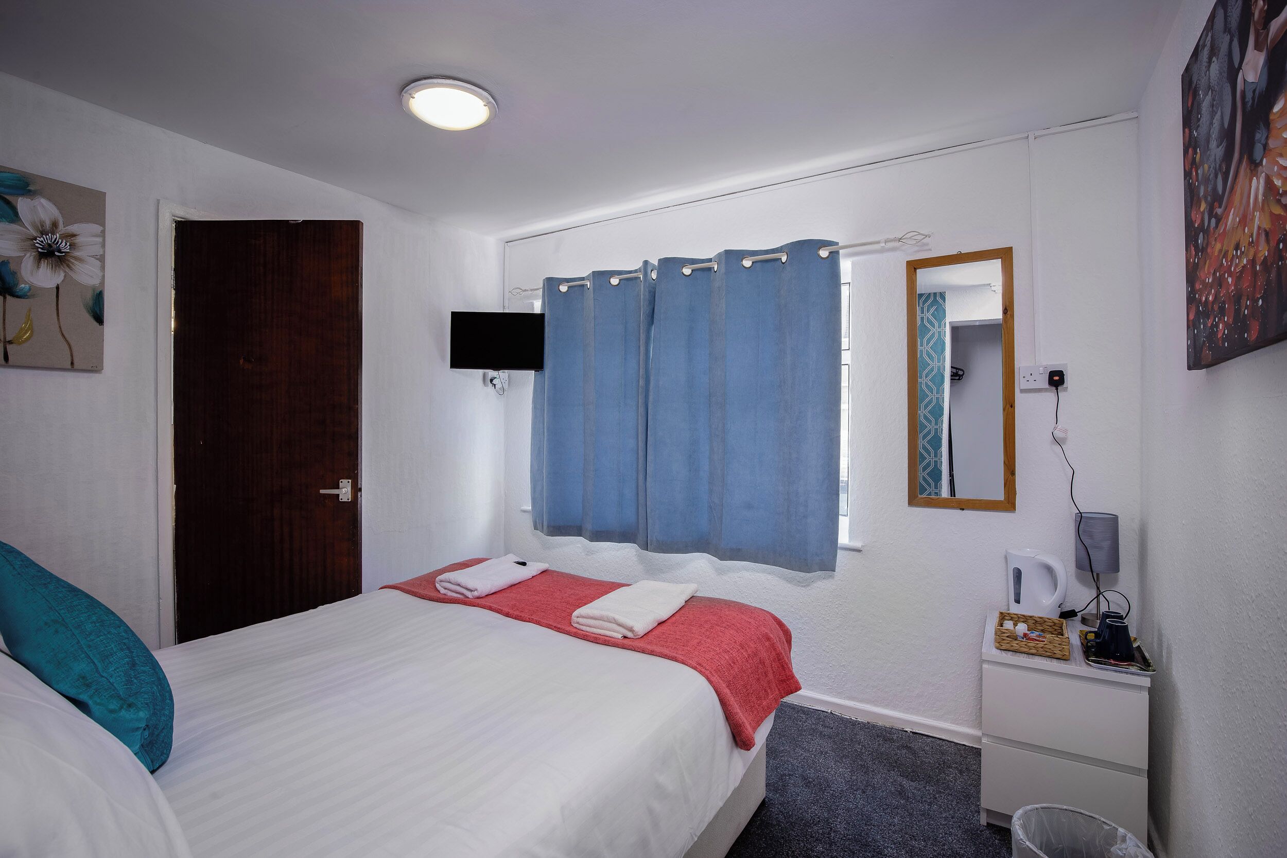 A hotel room with a bed, a desk with a kettle, a trash bin, and a window with blue curtains.
