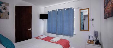 A hotel room with a bed, a desk with a kettle, a trash bin, and a window with blue curtains.