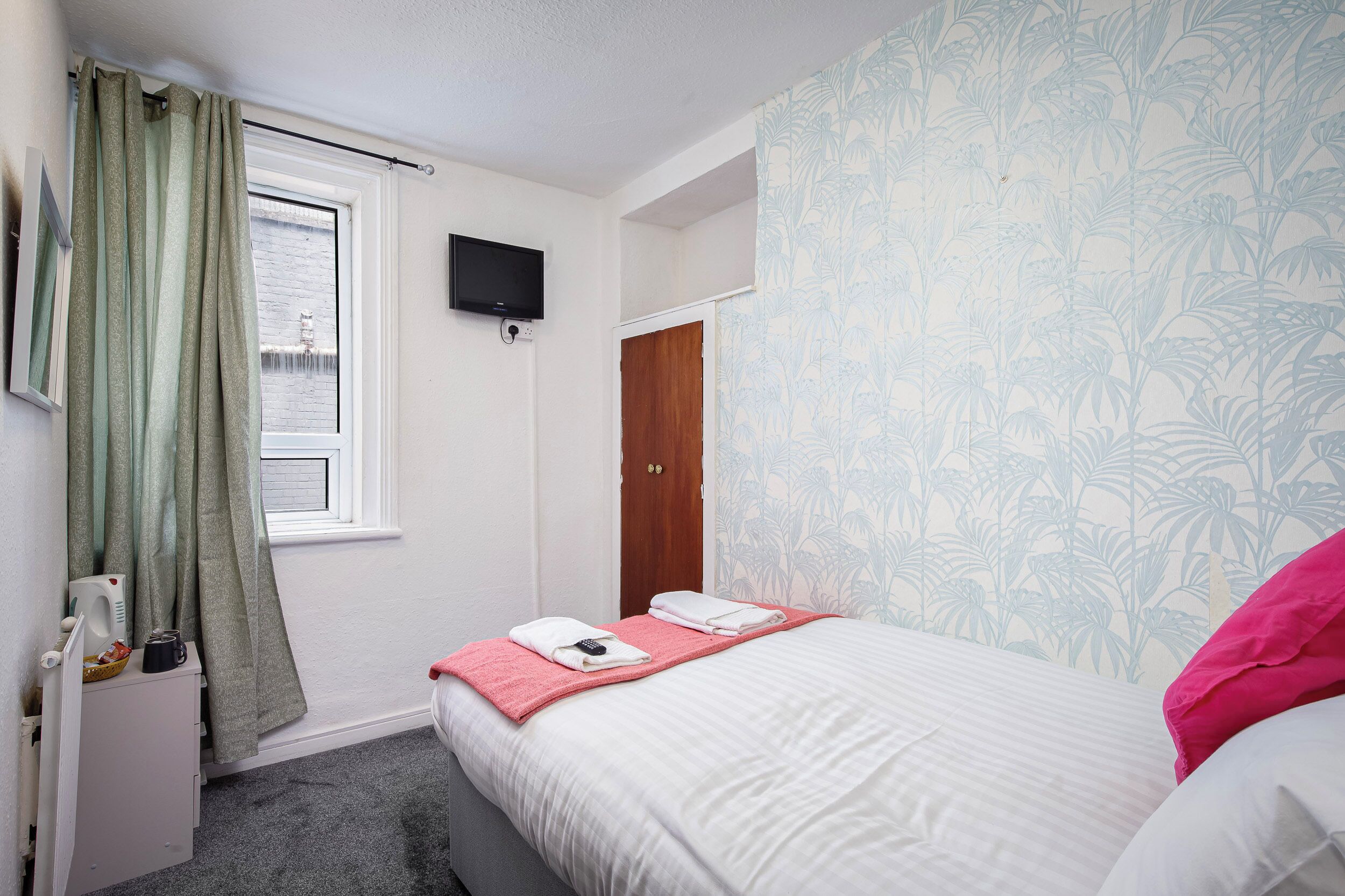 A hotel room with a bed, a television mounted on the wall, a window with curtains, and a door.