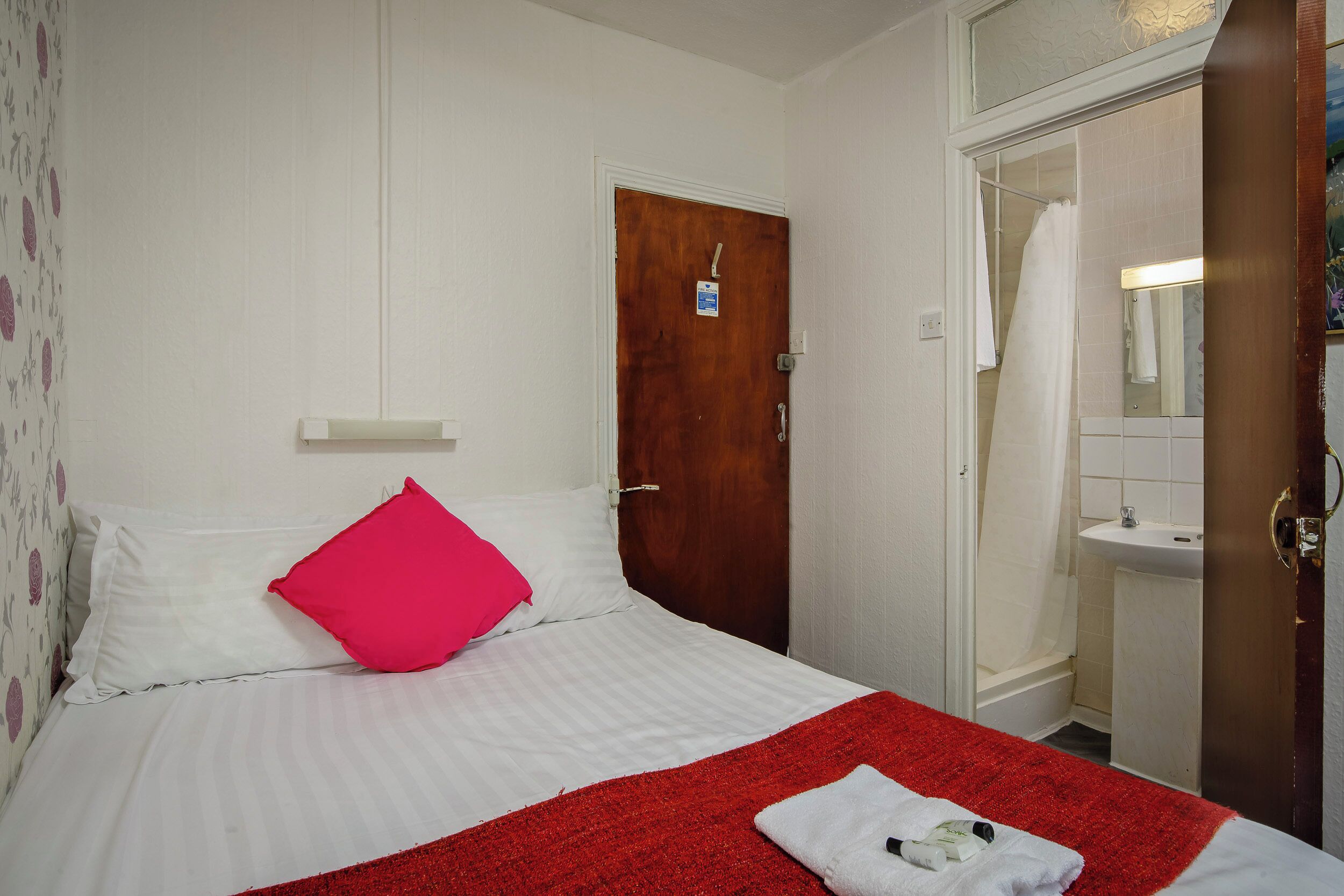 A hotel room with a bed, a red blanket, a white pillow, a wooden door, a bathroom with a sink and mirror, and a shower curtain.