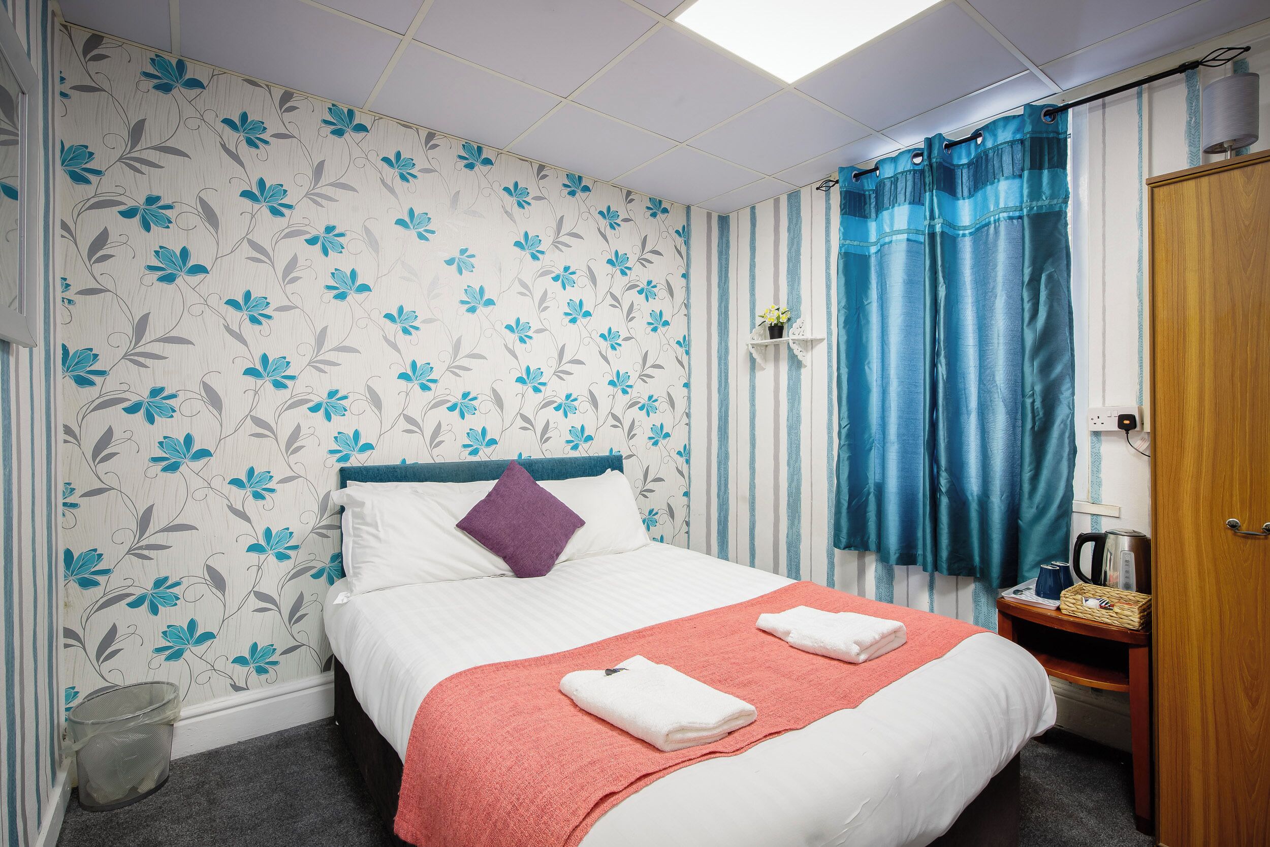 Een keurig opgemaakt bed met een rode deken, een paars kussen en twee gevouwen handdoeken. Er staat een klein houten nachtkastje met een waterkoker en een mandje. De kamer heeft een gestreept gordijn en bloemen behang.