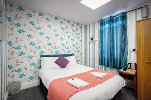 Una cama bien hecha con una manta roja, un cojín morado y dos toallas dobladas. Hay una mesita de noche de madera con una tetera y una cesta. La habitación tiene una cortina a rayas y un papel tapiz floral.