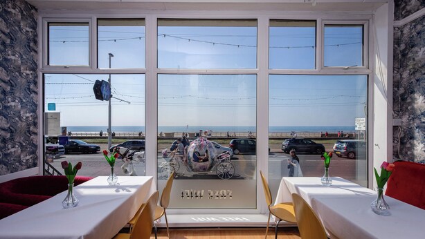 Een restaurant met grote ramen, met uitzicht op het strand en geparkeerde auto's.