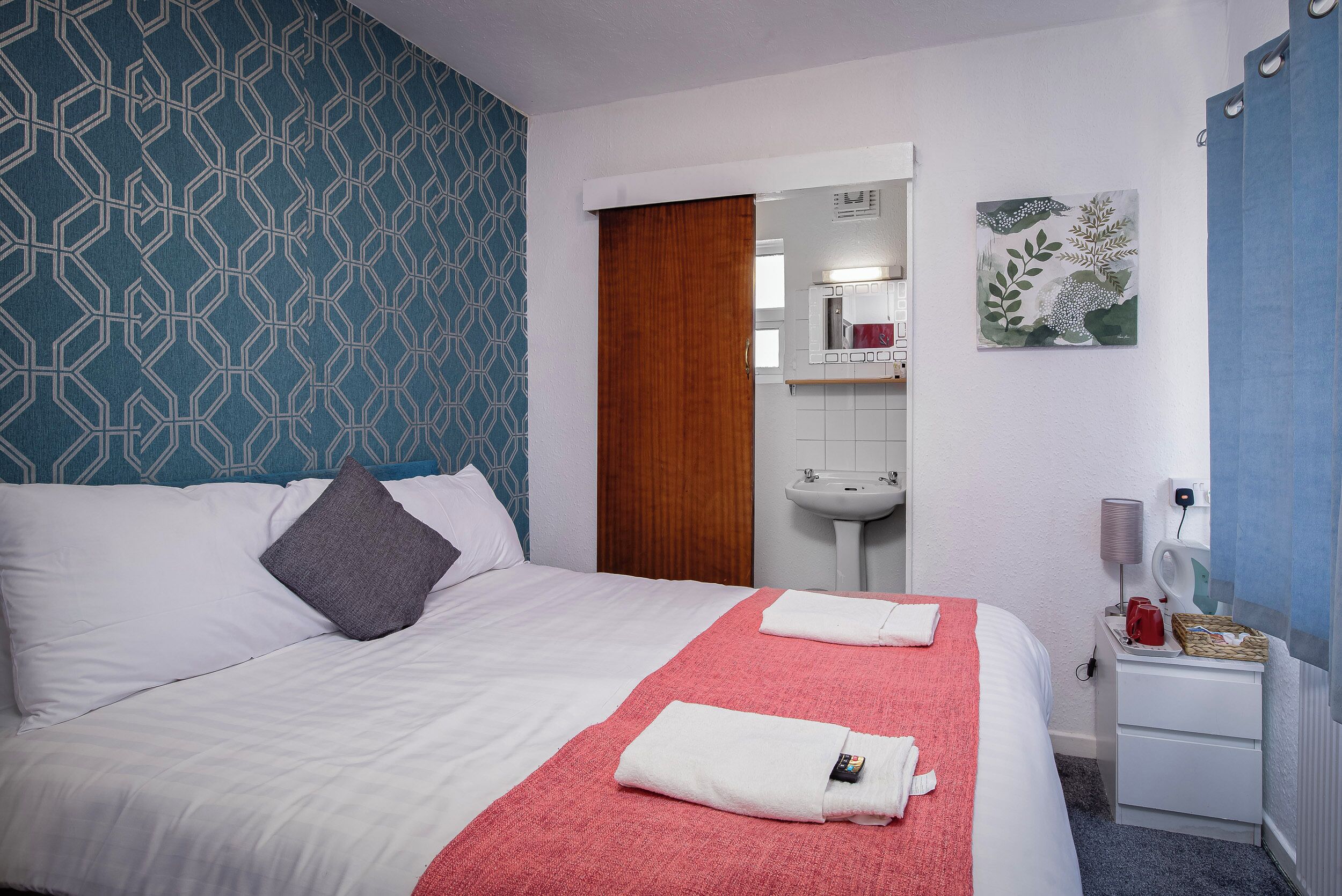 Habitación de hotel con una cama, una toalla roja, un almohadón blanco, una puerta de madera, un lavamanos y un espejo.