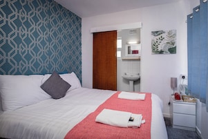 Habitación de hotel con una cama, una toalla roja, un almohadón blanco, una puerta de madera, un lavamanos y un espejo.