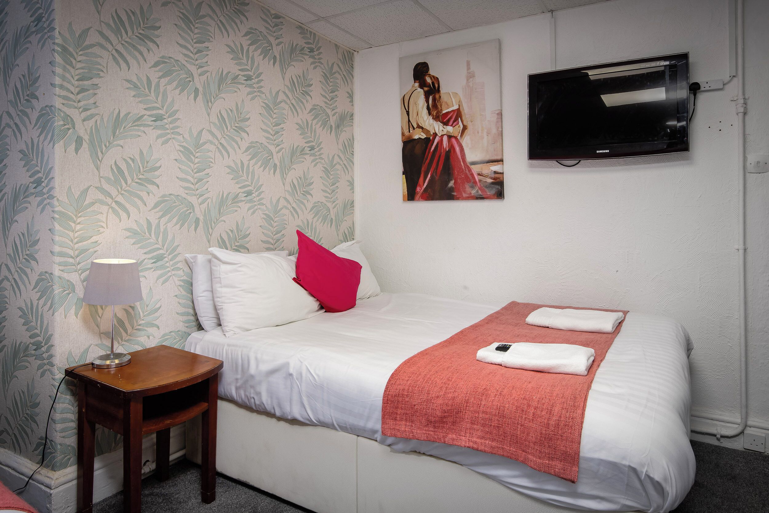 Un lit bien fait, avec du linge de lit blanc, un plaid rouge et deux serviettes pliées. Une petite table de chevet en bois avec une lampe se trouve à côté du lit. Un tableau représentant un couple est accroché au mur, au-dessus du lit.