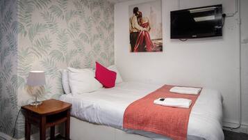 Een net opgemaakt bed met witte lakens, een rode plaid en twee gevouwen handdoeken. Een klein houten nachtkastje met een lamp staat naast het bed. Aan de muur boven het bed hangt een schilderij van een stel.