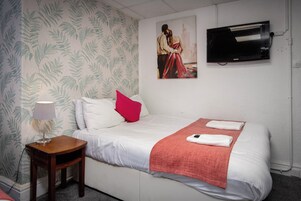 Een net opgemaakt bed met witte lakens, een rode plaid en twee gevouwen handdoeken. Een klein houten nachtkastje met een lamp staat naast het bed. Aan de muur boven het bed hangt een schilderij van een stel.