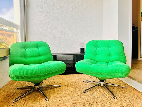 A room with two green upholstered chairs with metal legs, a wooden floor, a white textured wall, and a window with a view of buildings.