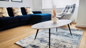 En stue med blå sofa, grå lænestol, hvidt sofabord og et tæppe med mønster.