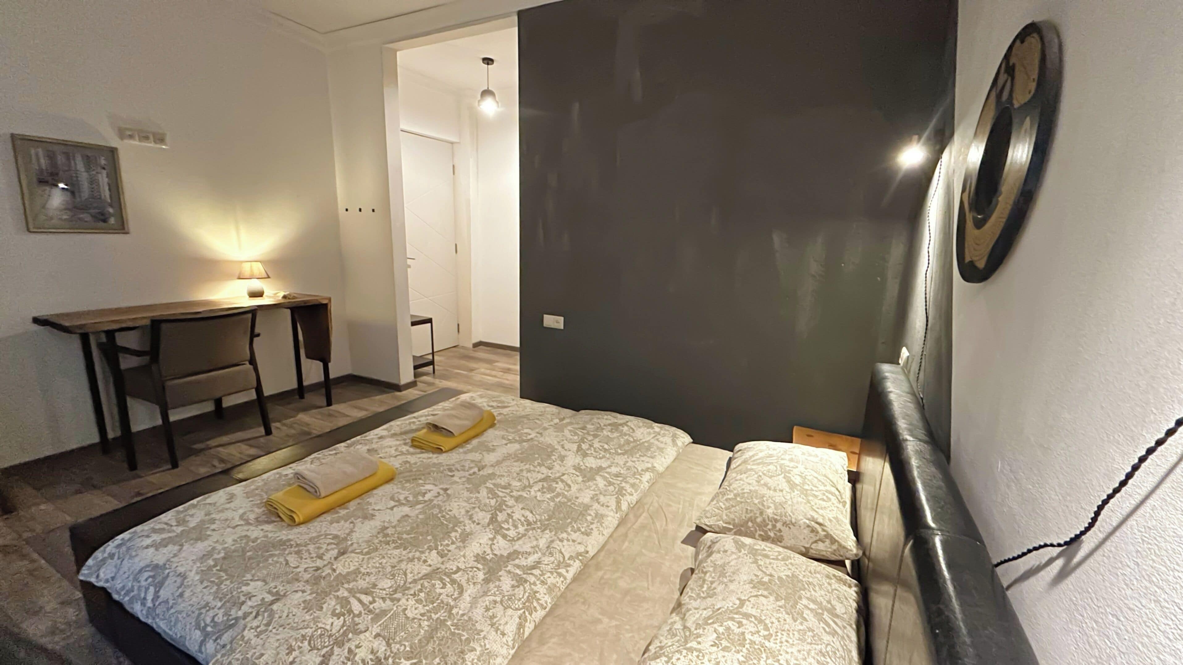 A hotel room with a bed, a desk, and a chair. There is a door leading to another room.