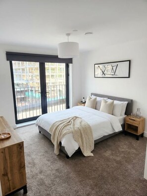 A modern bedroom with a large bed, a wooden nightstand, a bedside table, a hanging light fixture, and a view through a sliding glass door.