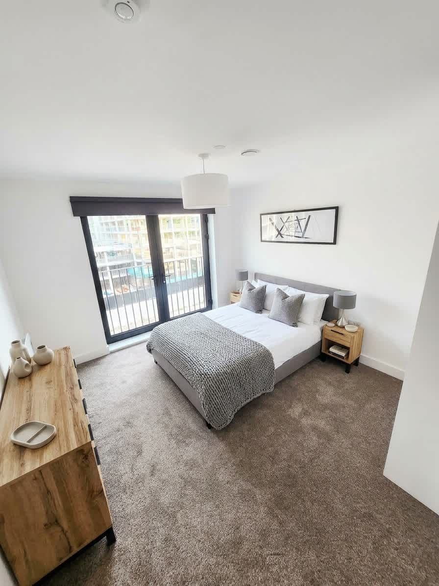 A bedroom with a bed, bedside tables, a wooden dresser, a large window, and a ceiling light.