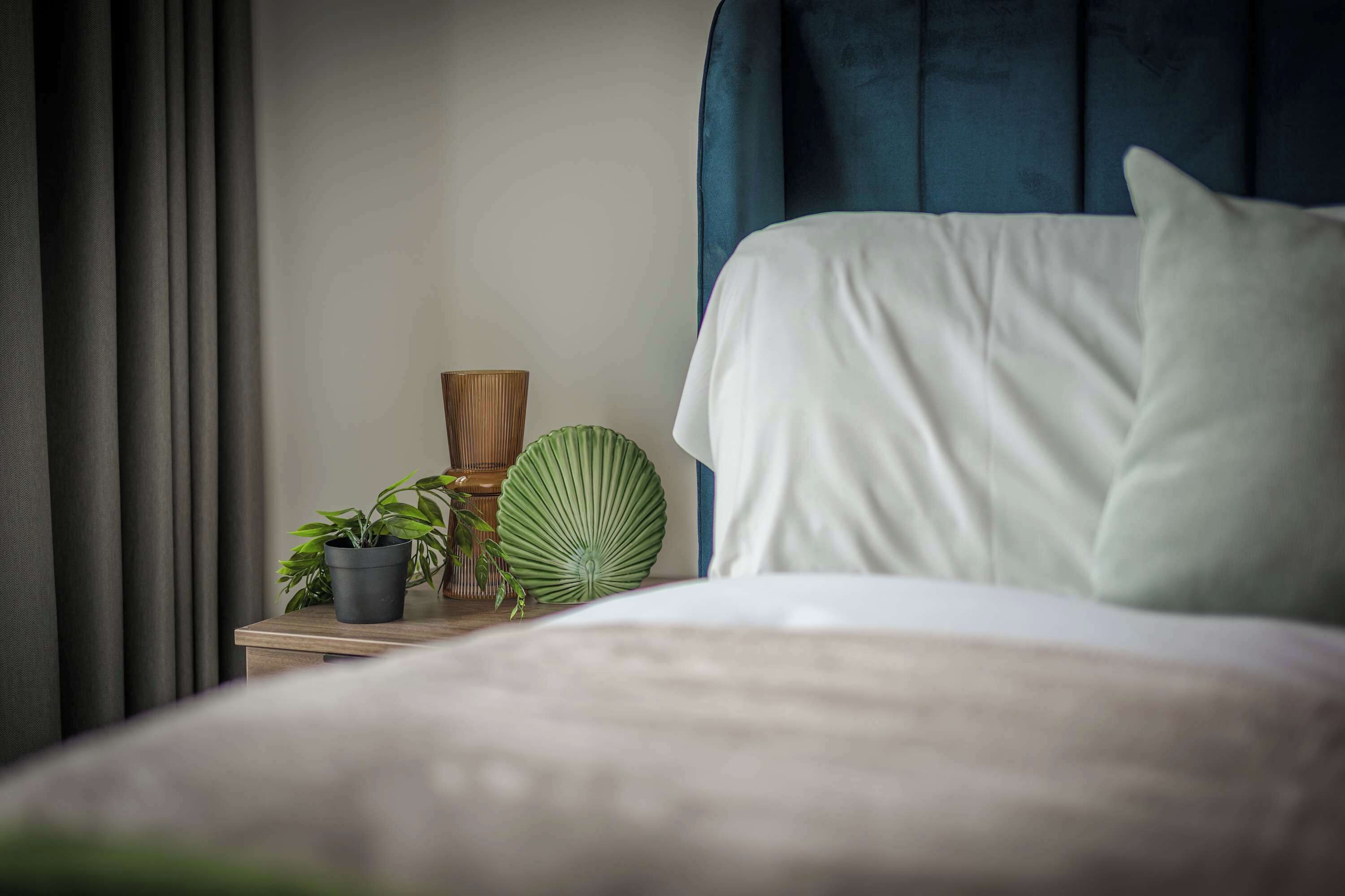 Un lit bien fait, avec une literie blanche, une tête de lit bleu foncé et une table de chevet agrémentée d’une plante en pot et d’objets décoratifs.