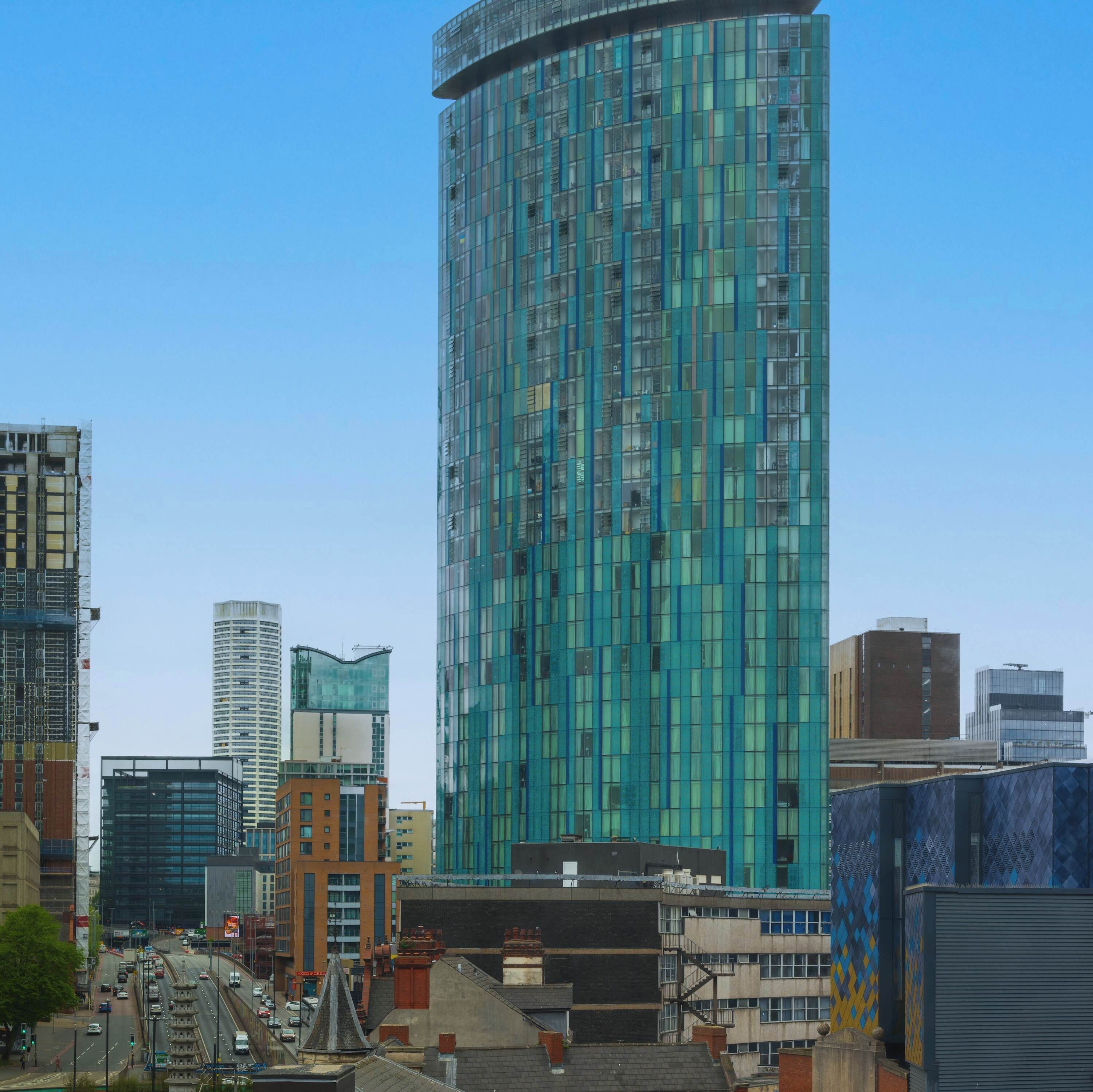 Un immeuble moderne de grande hauteur, doté d’une façade en verre, entouré d’autres bâtiments et bordé par une rue animée.