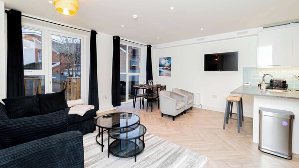 A modern living room with a black sofa, a glass coffee table, and a dining area with a table and chairs. There is a kitchen area with a sink and a TV mounted on the wall. - Stylish 2BR Stay in Tonbridge Town Centre (Tonbridge)