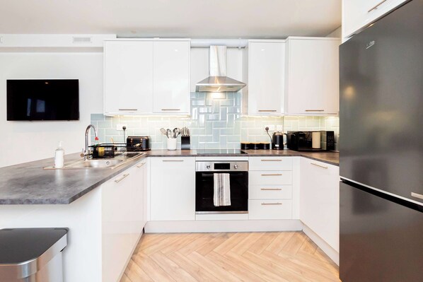 A modern kitchen with white cabinets, a stainless steel refrigerator, a built-in oven, and a flat-screen TV mounted on the wall. - Stylish 2BR Stay in Tonbridge Town Centre (Tonbridge)