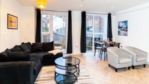 A modern living room with a black sofa, a glass coffee table, and a white armchair. There is a dining area with a table and chairs visible through large windows. - Stylish 2BR Stay in Tonbridge Town Centre (Tonbridge)