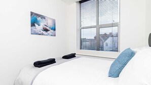 A hotel room with a bed, a painting of a boat on water, and a window with blinds. - Stylish 2BR Stay in Tonbridge Town Centre (Tonbridge)
