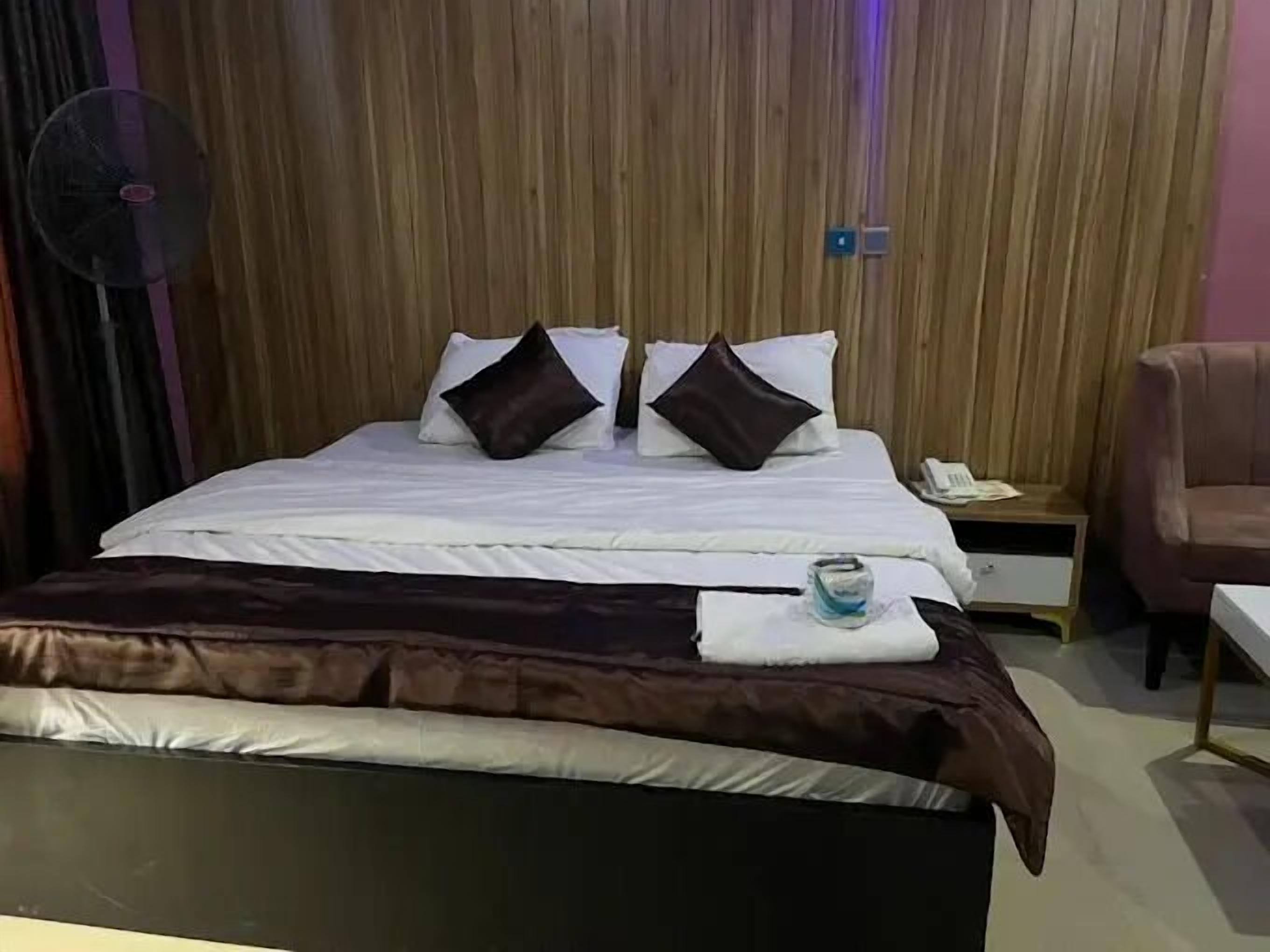 A neatly made bed with white and brown bedding, a bedside table with a phone and papers, and a fan on the left.