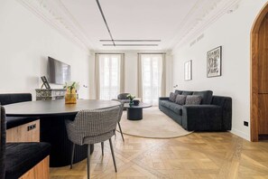 A modern living room with a dark grey sofa, a round dining table with chairs, and a large window with curtains.