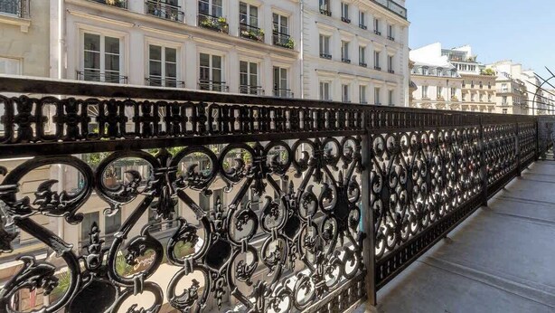 A wrought iron fence with circular mirrors, white buildings with balconies, and a clear sky. - ONIRI - Serviced 2-BR/2BA - Louvre Richelieu II (Paris)