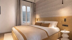 A modern hotel room with a large bed, a bedside table, a window with curtains, and a wall-mounted light fixture.
