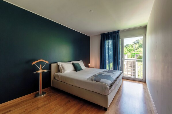 A bedroom with a bed, a bedside table, a floor lamp, and a view of the outdoors through a window.