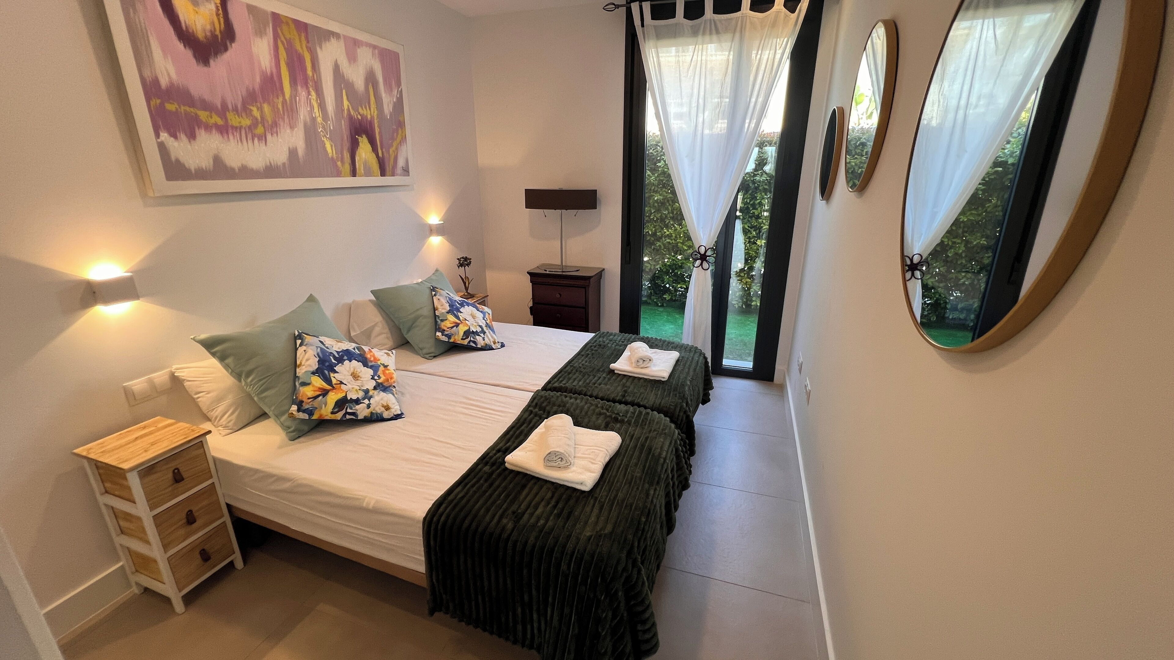 A bedroom with two beds, a bedside table, a lamp, a mirror, and a painting on the wall.