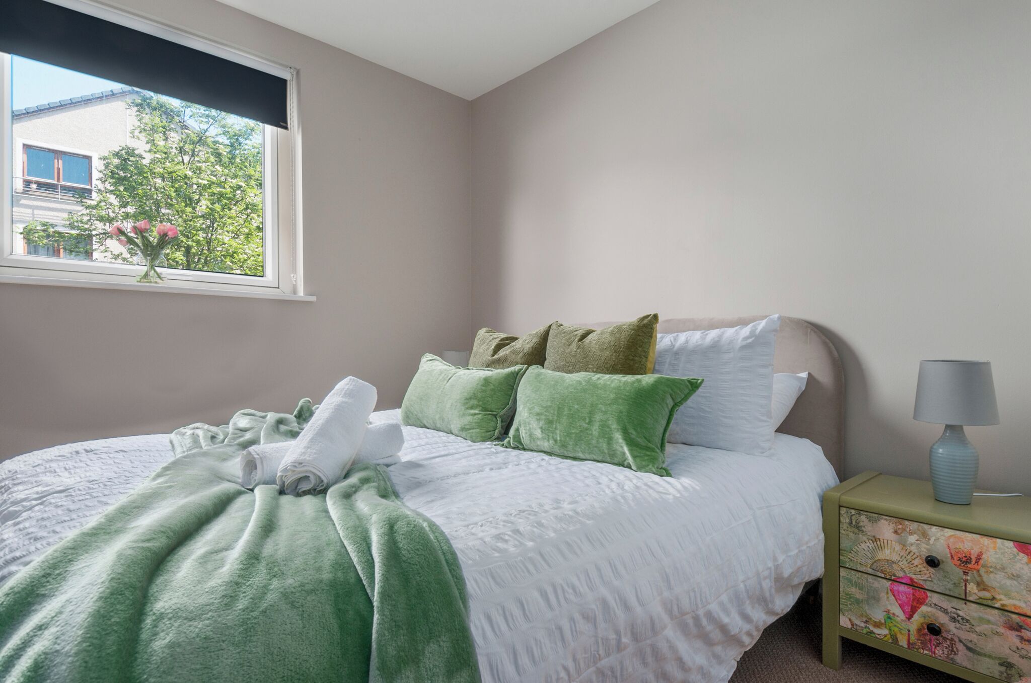 Una cama bien hecha con ropa de cama verde y blanca, una mesita de noche con una lámpara y una ventana con vistas a un edificio y árboles.