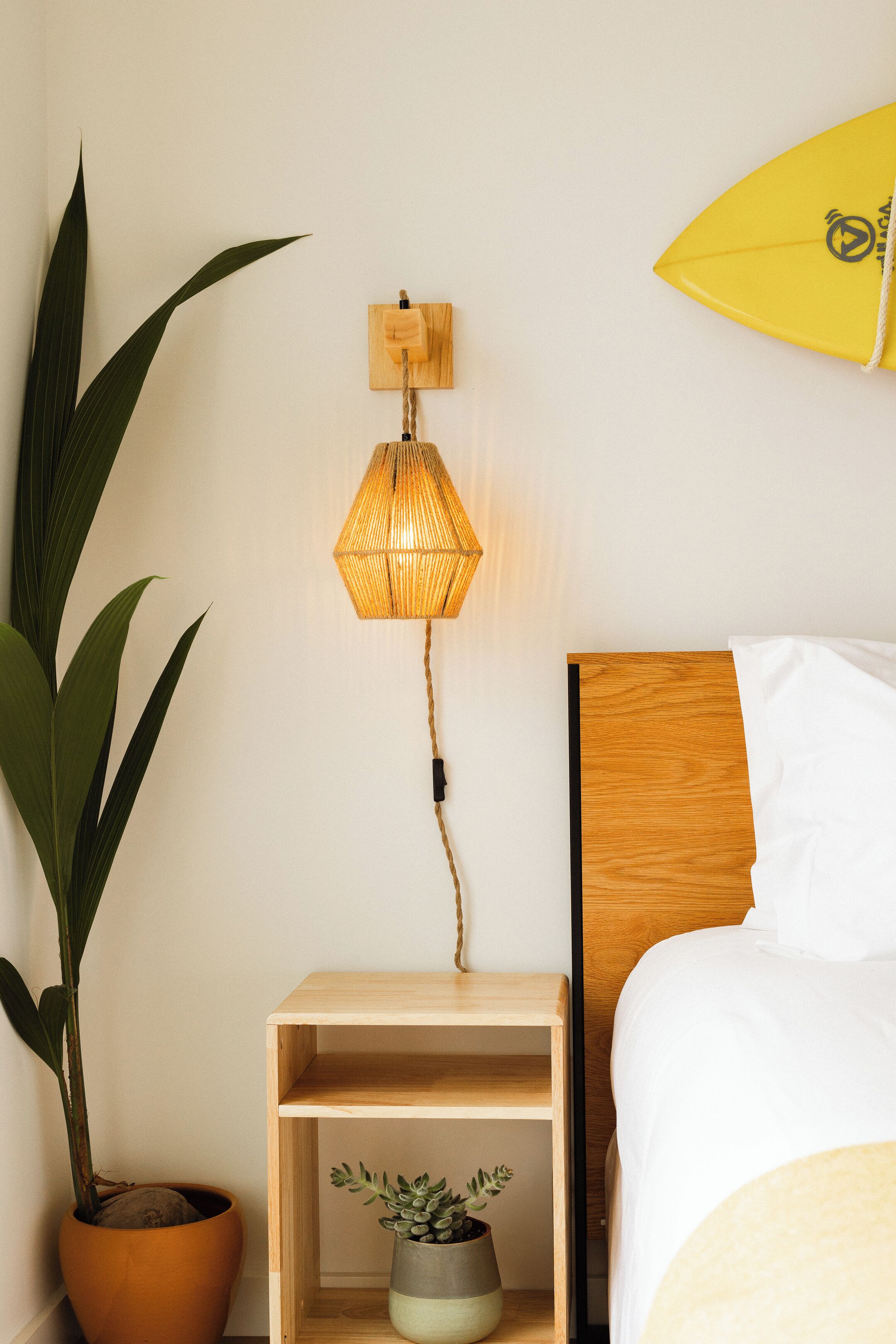 Une chambre d’hôtel bien rangée, avec une table de chevet en bois, une plante en pot, une lampe fixée au mur et un lit recouvert de draps blancs.