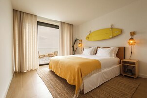 Habitación de hotel con cama, una tabla de surf amarilla en la pared, ventana con cortinas y una mesita de noche con lámpara.